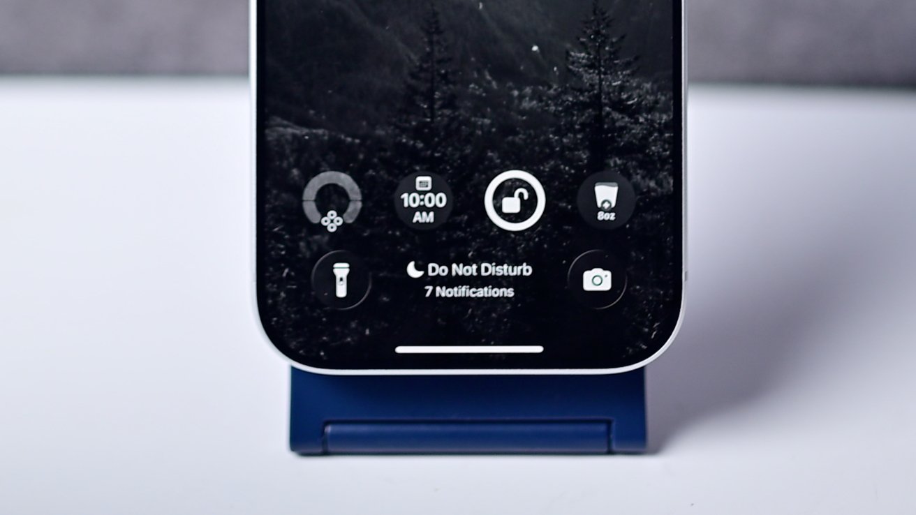 Smartphone on a blue stand displaying lock screen with time, weather, Do Not Disturb mode, and notifications. Background shows dark forest scenery.