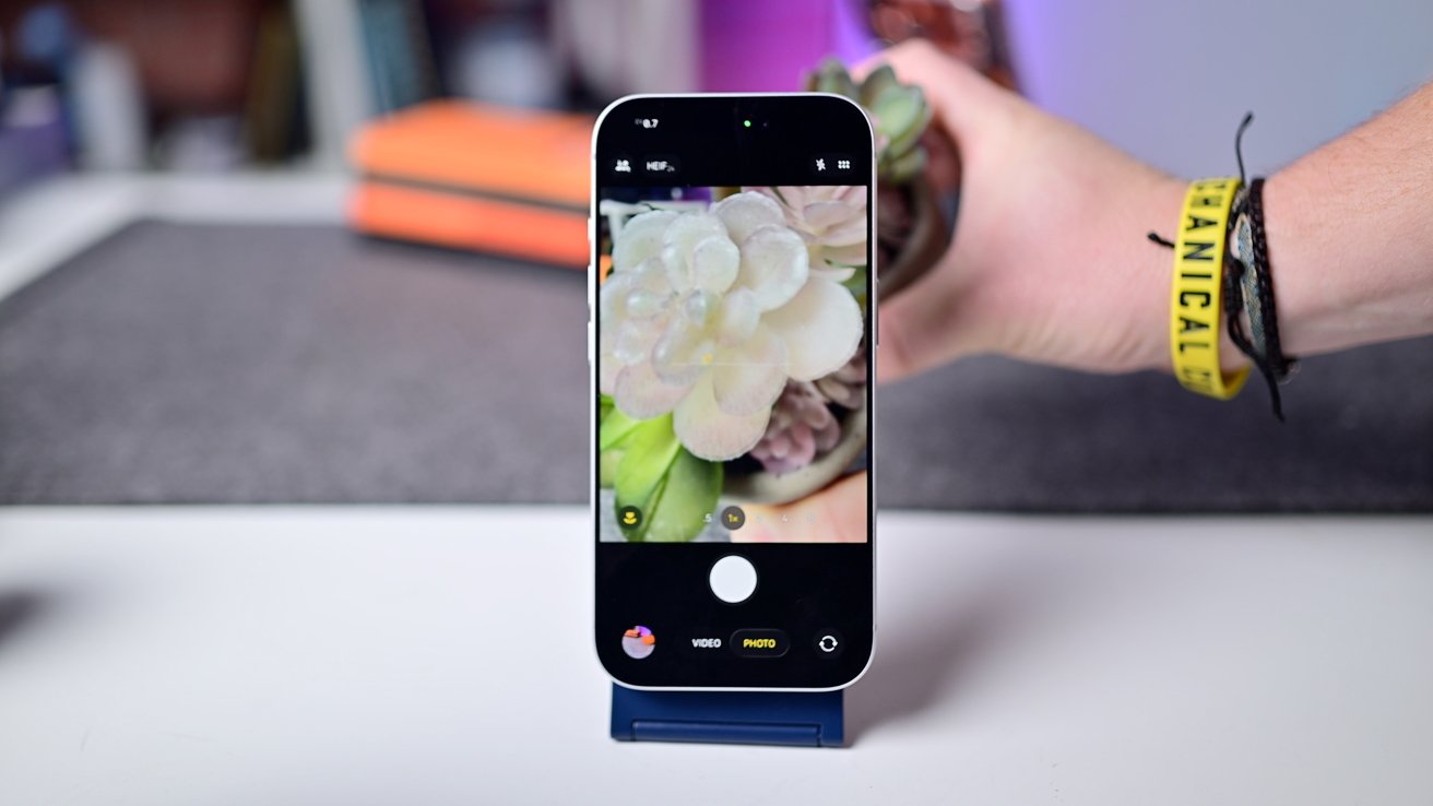 Smartphone capturing a close-up photo of a succulent plant. Hand wearing bracelets holding the plant, with a blurred background of a desk and orange notebook.