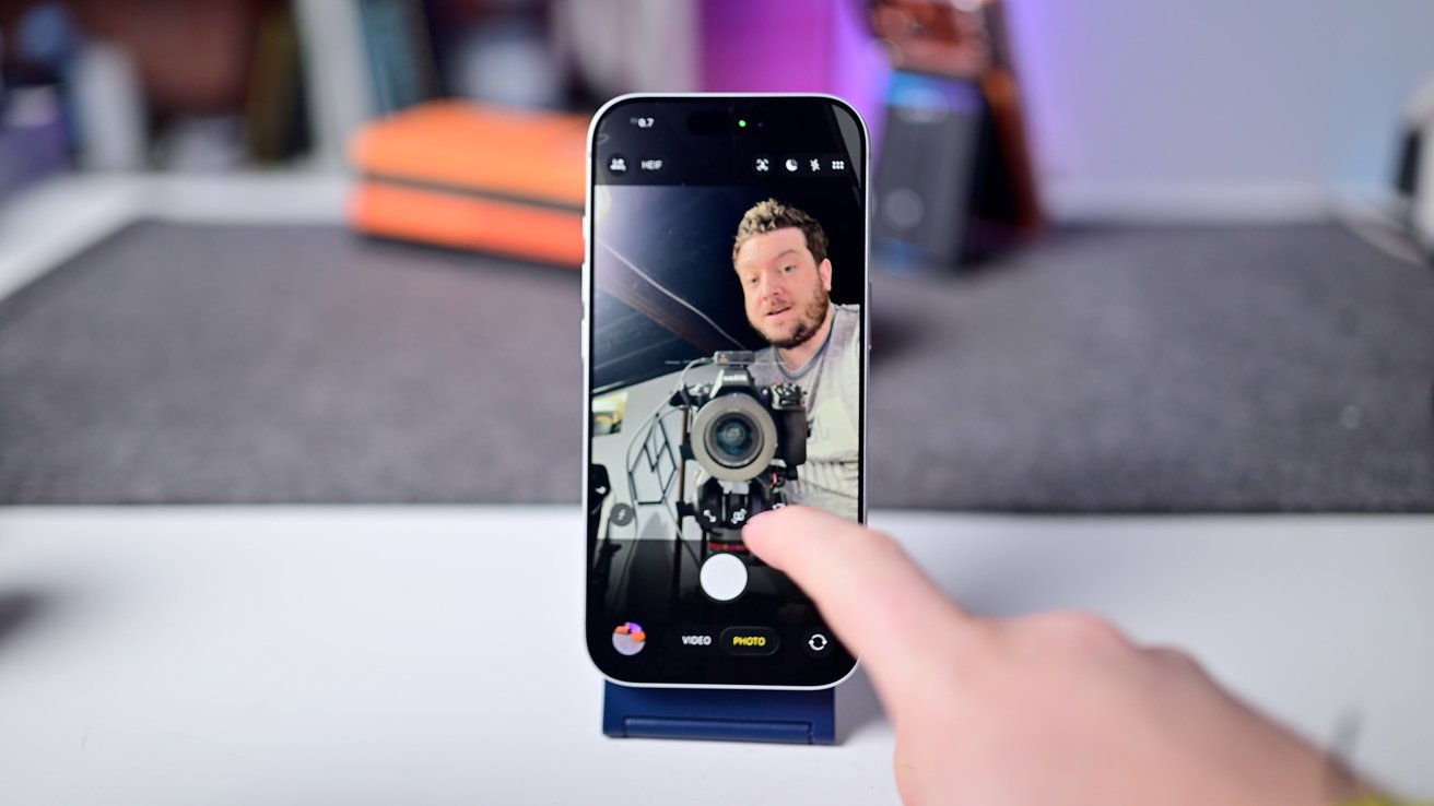 Person behind a camera is visible on a smartphone screen showing a selfie view, with a hand touching the screen.