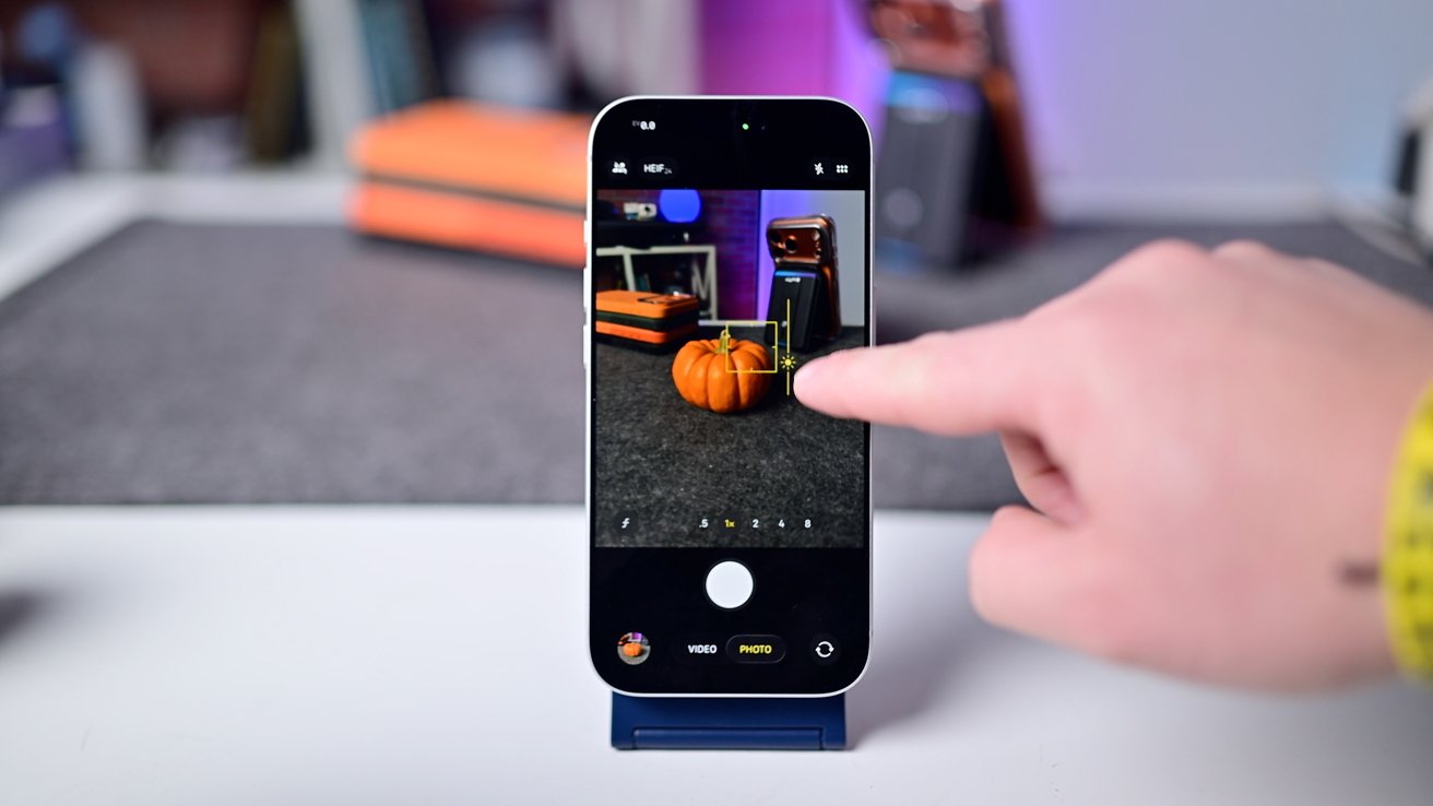A person interacts with a smartphone camera app, focusing on a small pumpkin on a table. The scene includes additional blurred objects in the background.