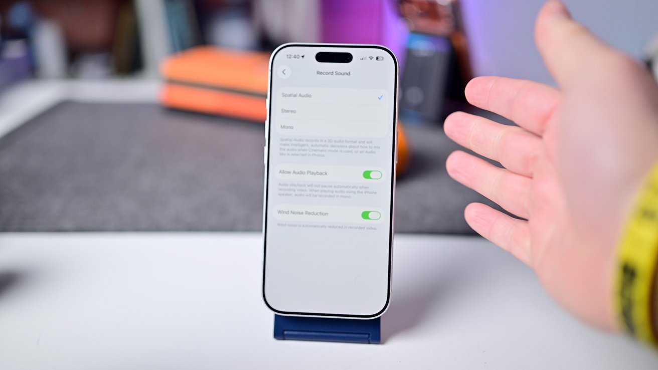Smartphone on a stand displaying audio settings screen, with a hand gesturing next to it, blurred background showing various desk items.