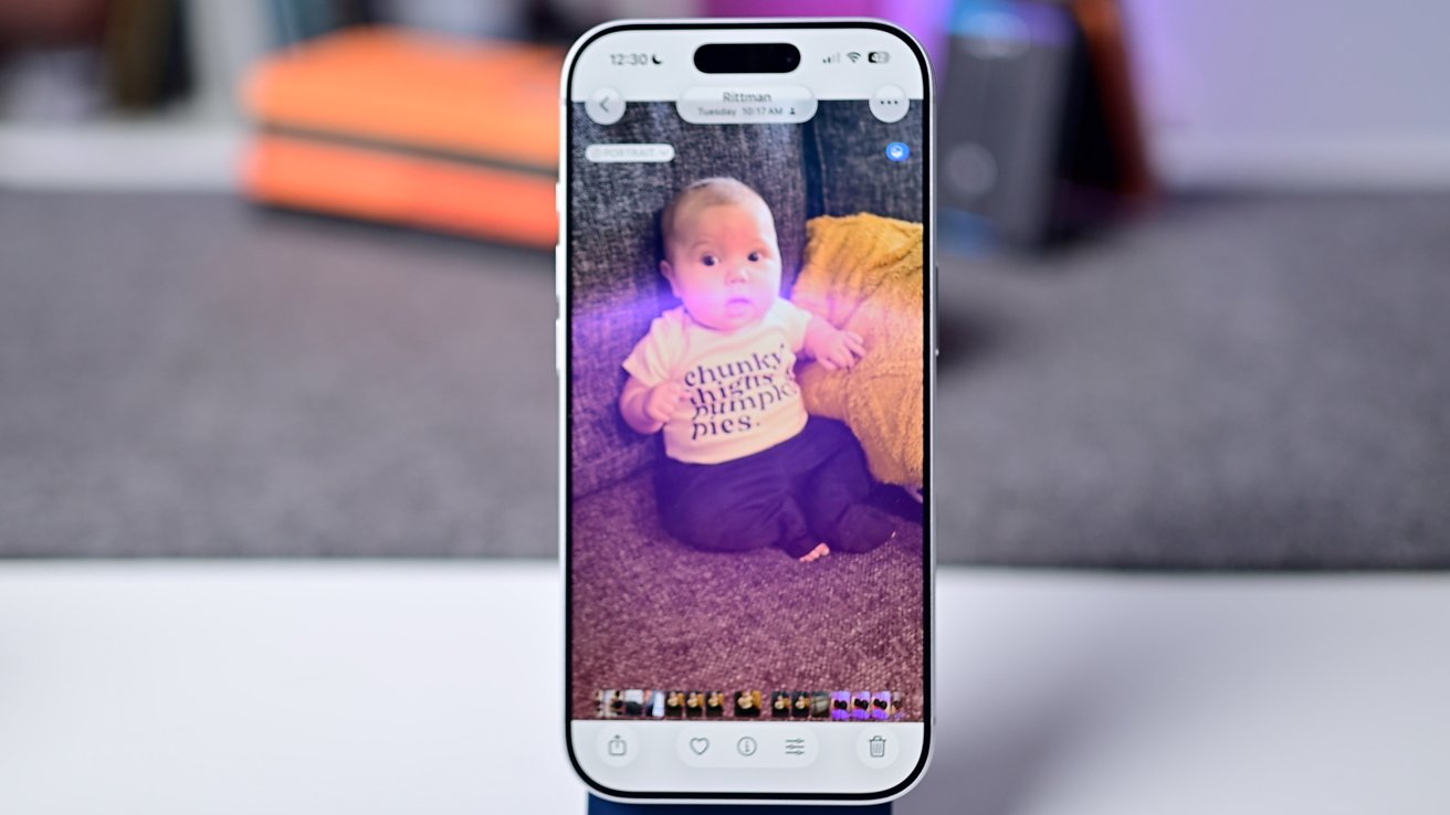 Smartphone displaying a baby wearing a printed shirt, sitting on a couch with a yellow pillow. A blurred background shows books and a speaker.