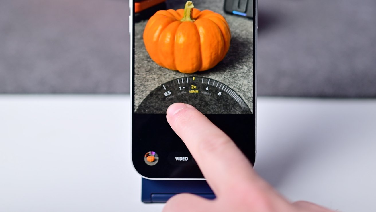 Finger touching smartphone screen displaying a small orange pumpkin with a zoom interface at 2x magnification.