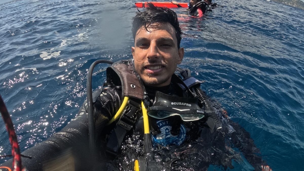 A scuba diver smiling in the ocean, wearing scuba gear with a second diver in the background and sunlight reflecting on the water.