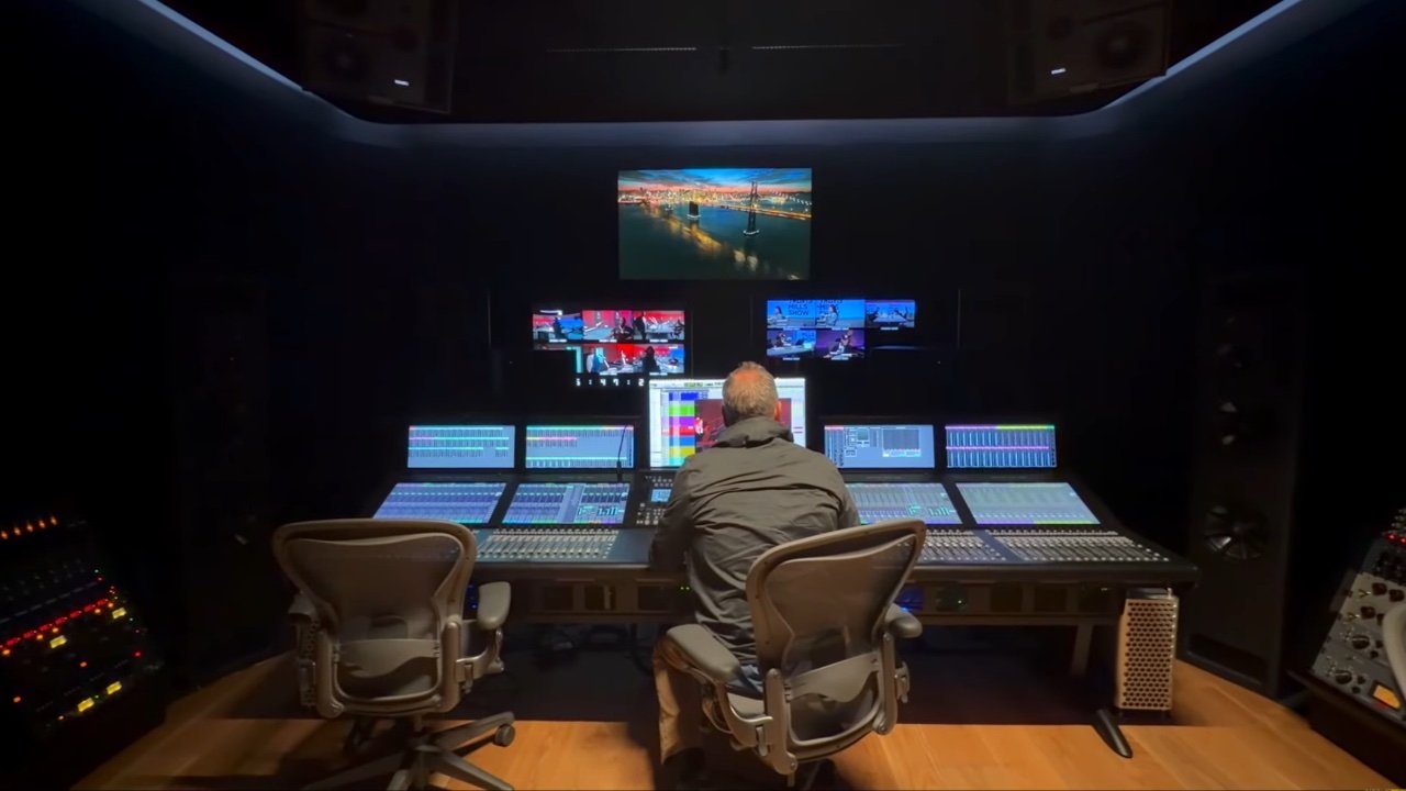 Person seated in a large, dimly lit recording studio with multiple screens and audio equipment, focusing on controls and displays.