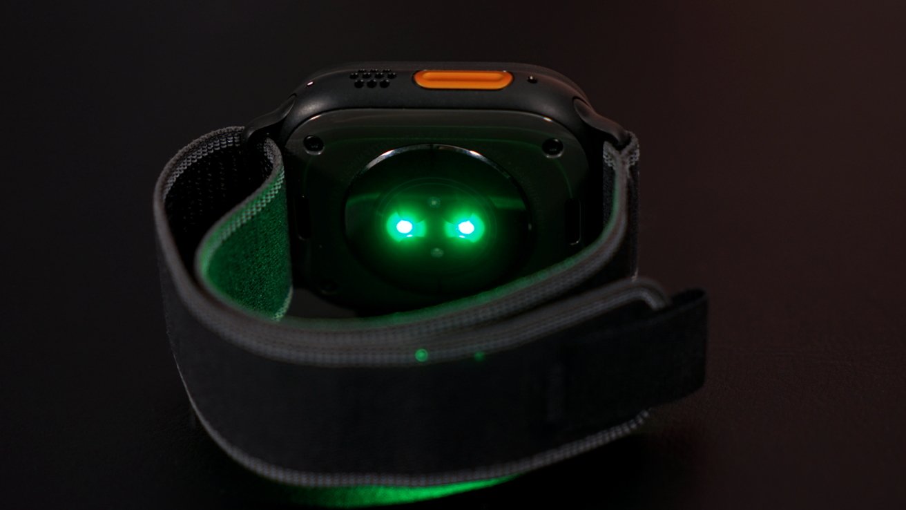 A smartwatch lying face-down with green lights glowing from the back, on a dark surface.