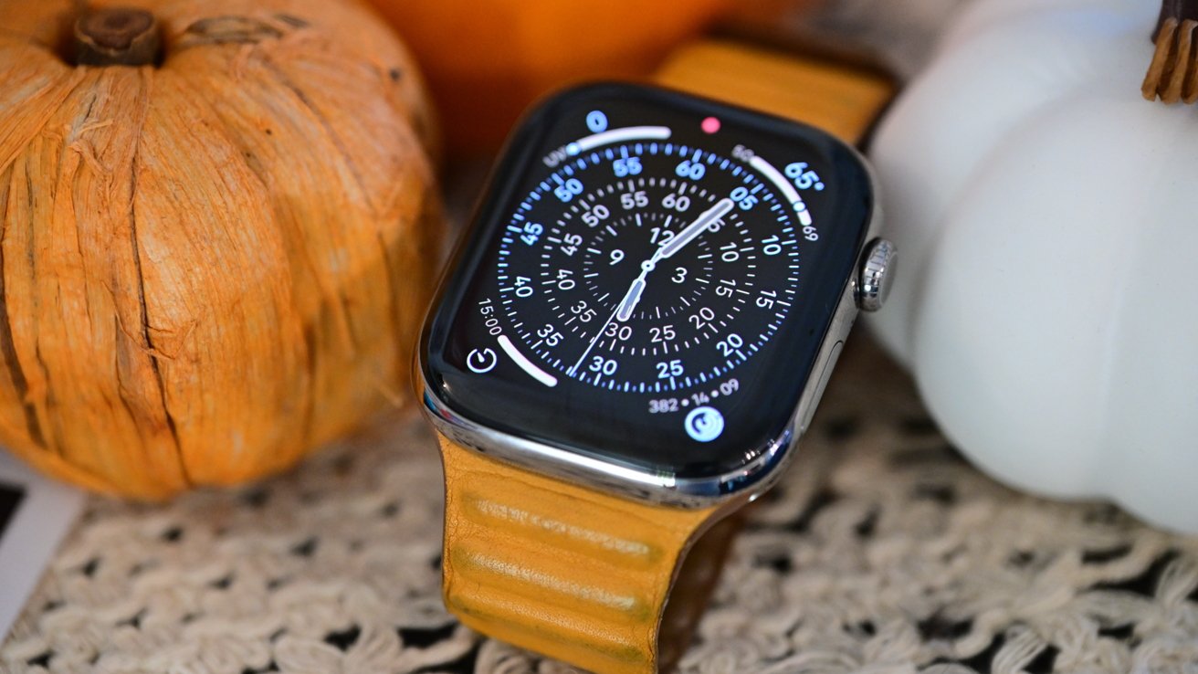 Smartwatch with orange leather strap displays analog time, surrounded by a large orange pumpkin and a smaller white pumpkin on a textured surface.