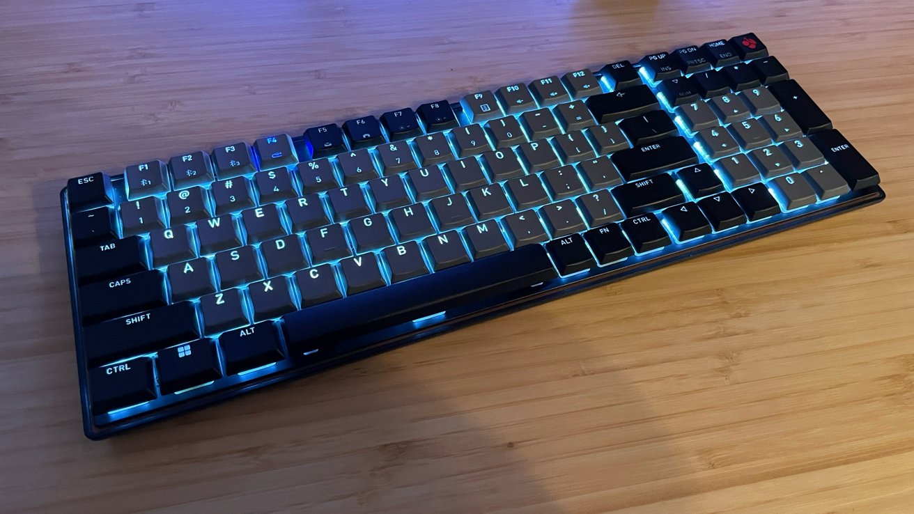 A mechanical keyboard with blue backlighting, keys arranged in a standard layout, sits on a wooden surface.