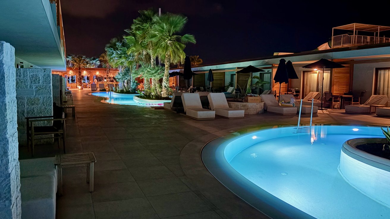 Nighttime poolside scene with illuminated water, palm trees, lounge chairs, and umbrellas.