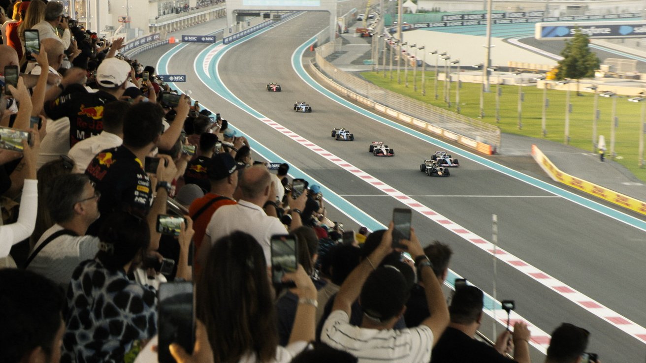 Crowd watches a car race on a track, phones raised, capturing the fast-moving cars with enthusiasm.