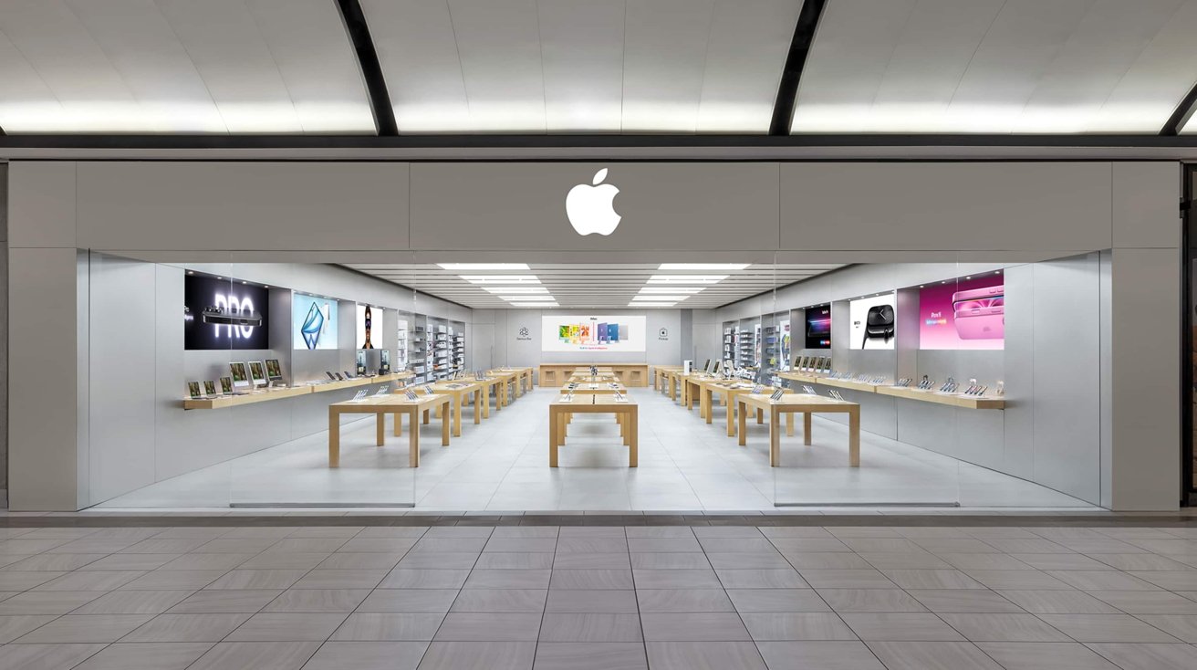 Modern Apple store interior with sleek wooden tables, electronic devices on display, illuminated white apple logo, and colorful marketing posters on the walls.