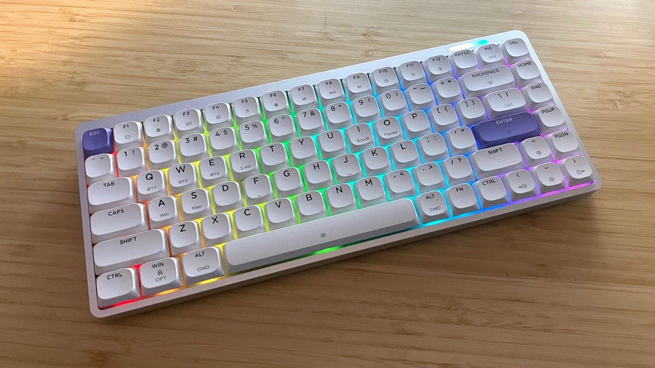 White compact keyboard with colorful backlighting, featuring purple escape and enter keys, on a light wood surface.
