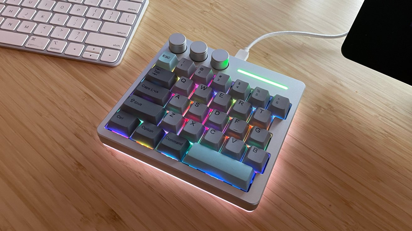 Compact mechanical keyboard with colorful backlighting and three silver dials on a wooden desk, next to an Apple keyboard.