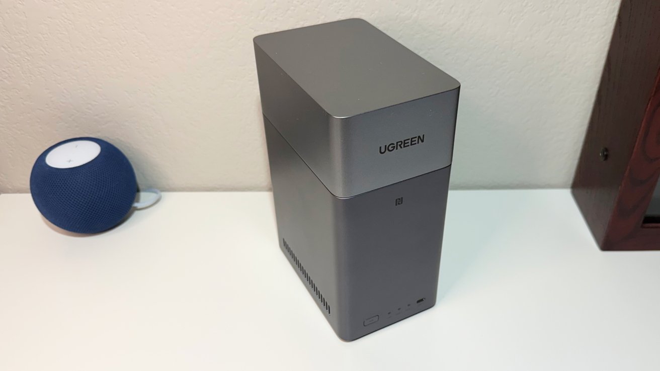 Gray vertical rectangular speaker with 'UGREEN' branding beside a small blue spherical speaker on a white surface.
