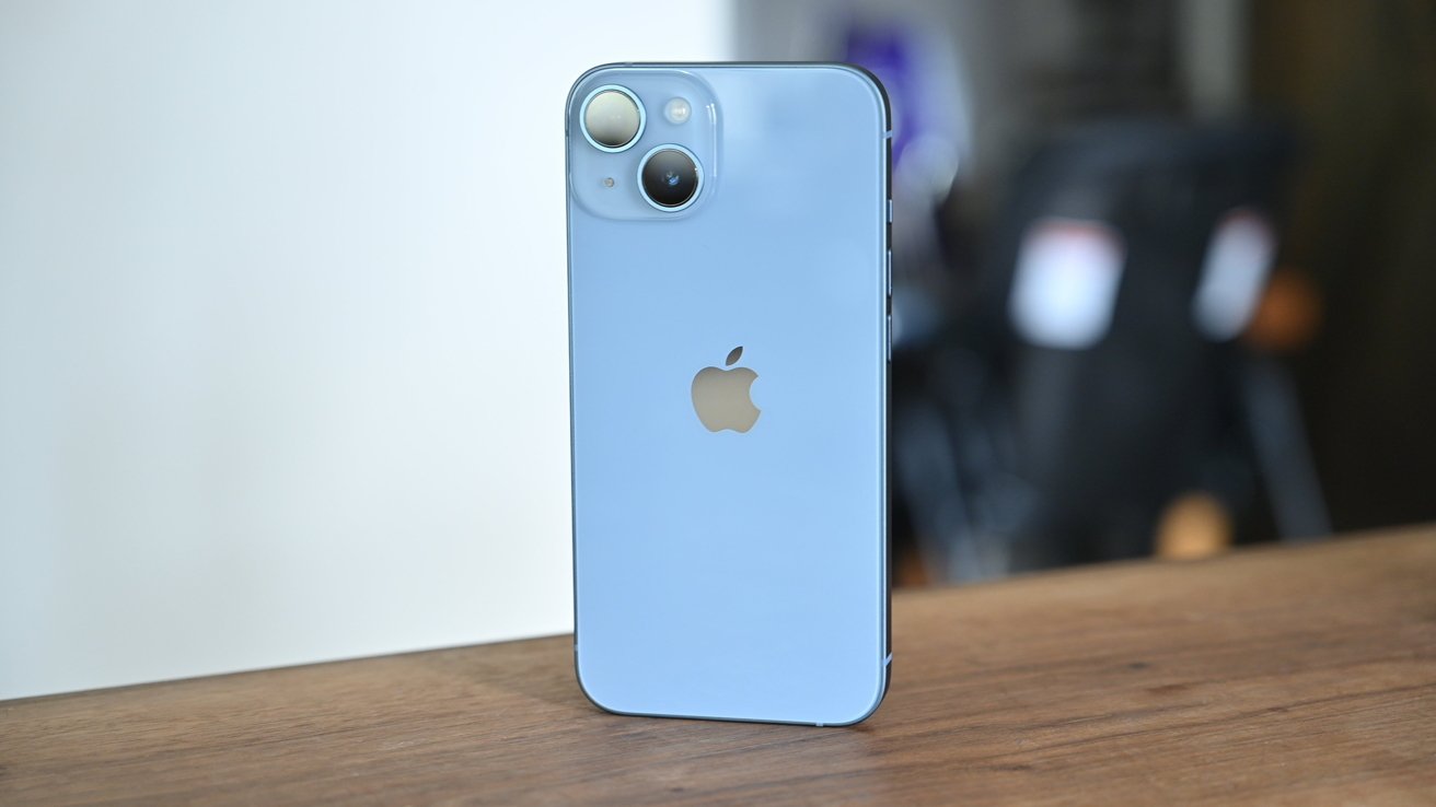 Blue smartphone with dual cameras and Apple logo stands on a wooden surface, blurred background.