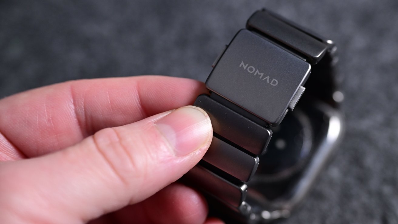 Fingers holding a dark metal watch band with the word 'NOMAD' on the clasp, blurred background.