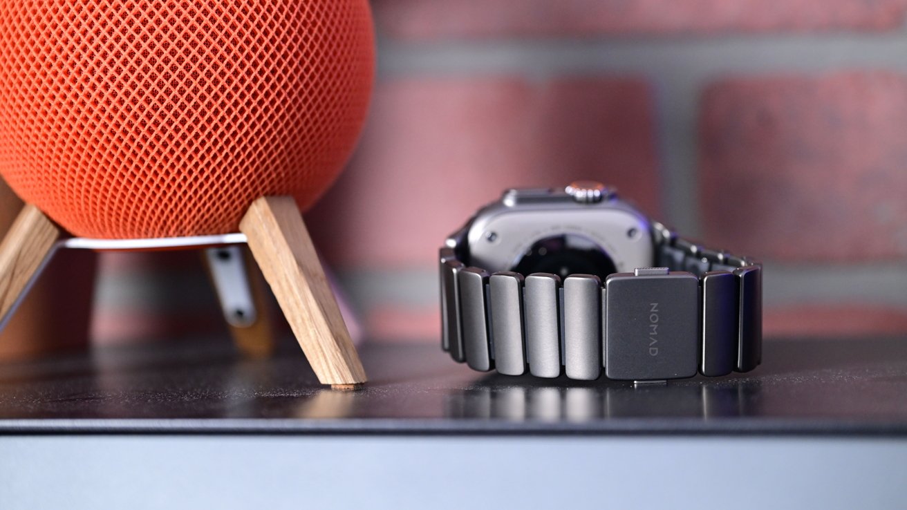 An orange mesh ball on a wooden stand next to a metal watch with a black segment band, against a blurred brick background.