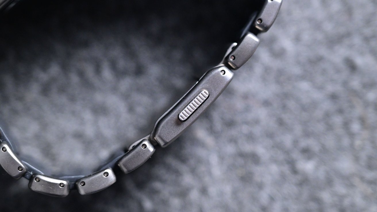 Close-up of a metal watch bracelet with visible links and a textured clasp on a blurred gray background.