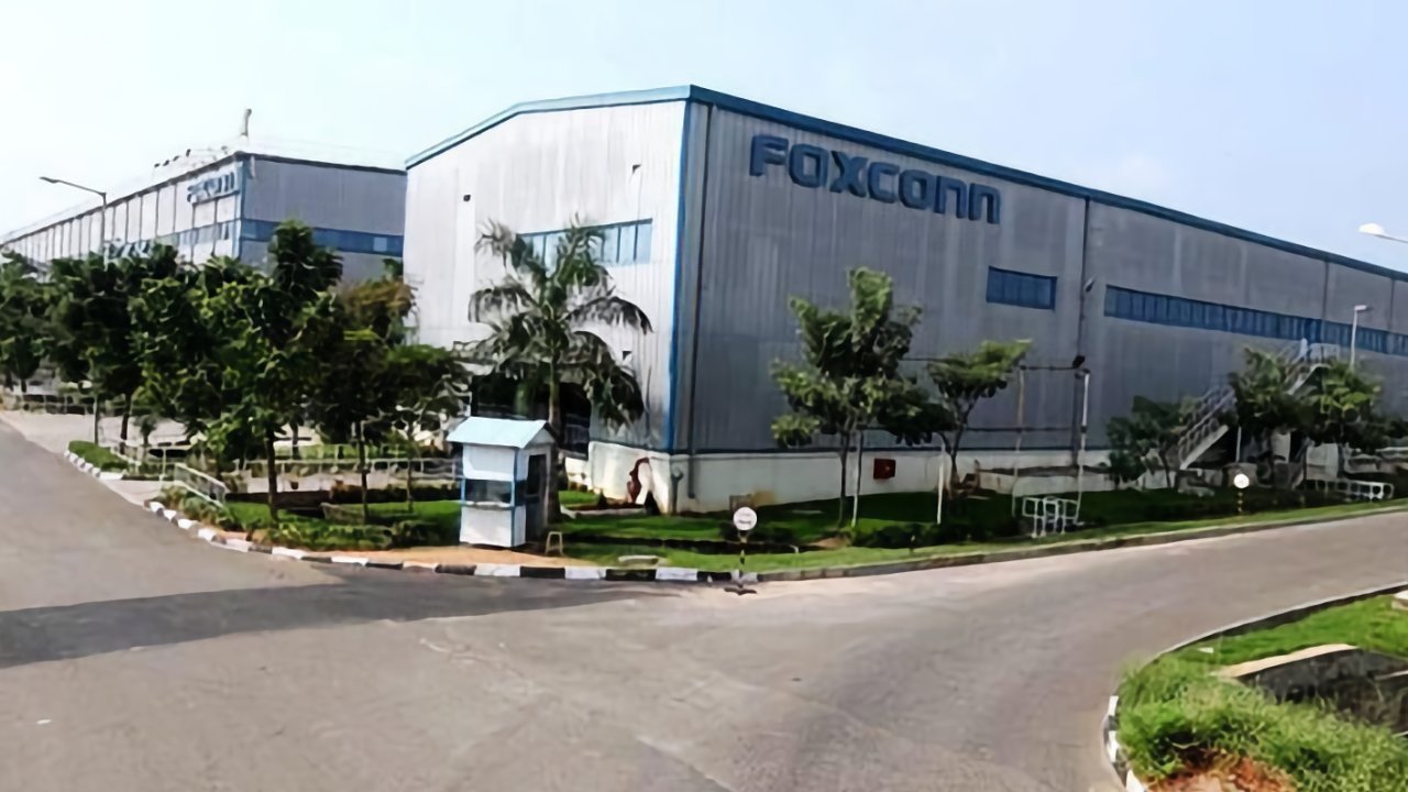 Large industrial building with Foxconn logo, surrounded by greenery and trees, situated at an intersection with a small security booth nearby.