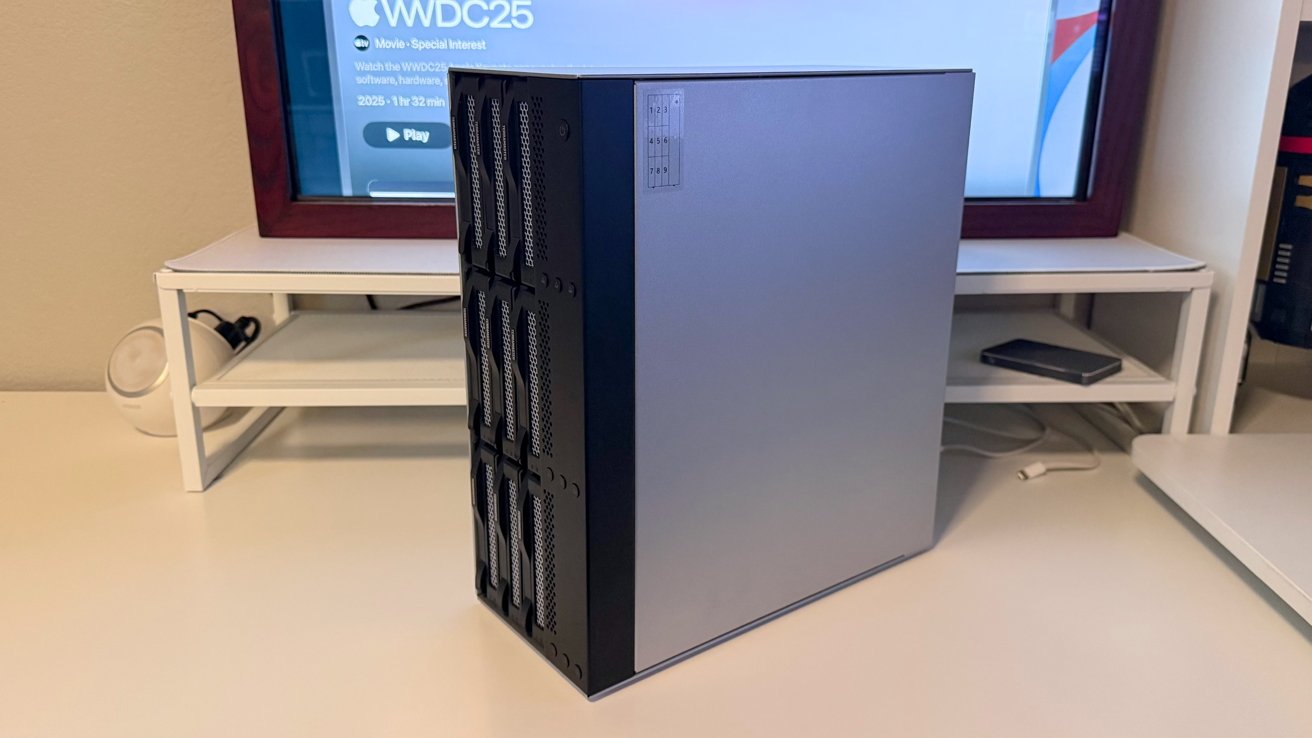 A silver desktop computer tower with ventilation slots on a white desk, TV screen and shelf with a smartphone in the background.