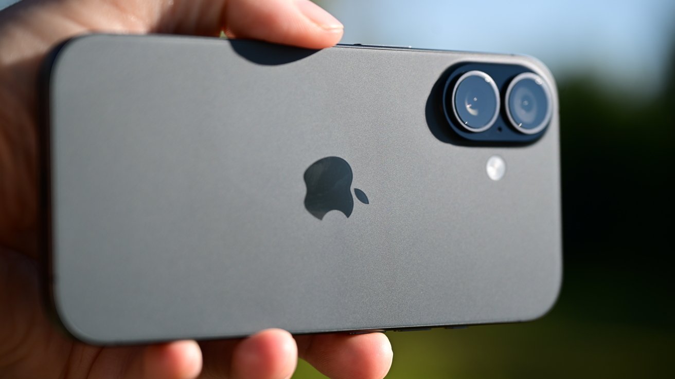A person holds a sleek, modern smartphone with a dual camera and Apple logo, against a blurred outdoor background.