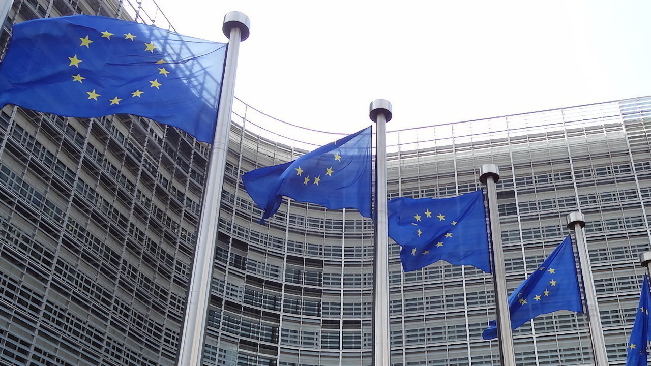 Five blue flags with yellow stars arranged in a circle, flying in front of a modern, curved glass building.