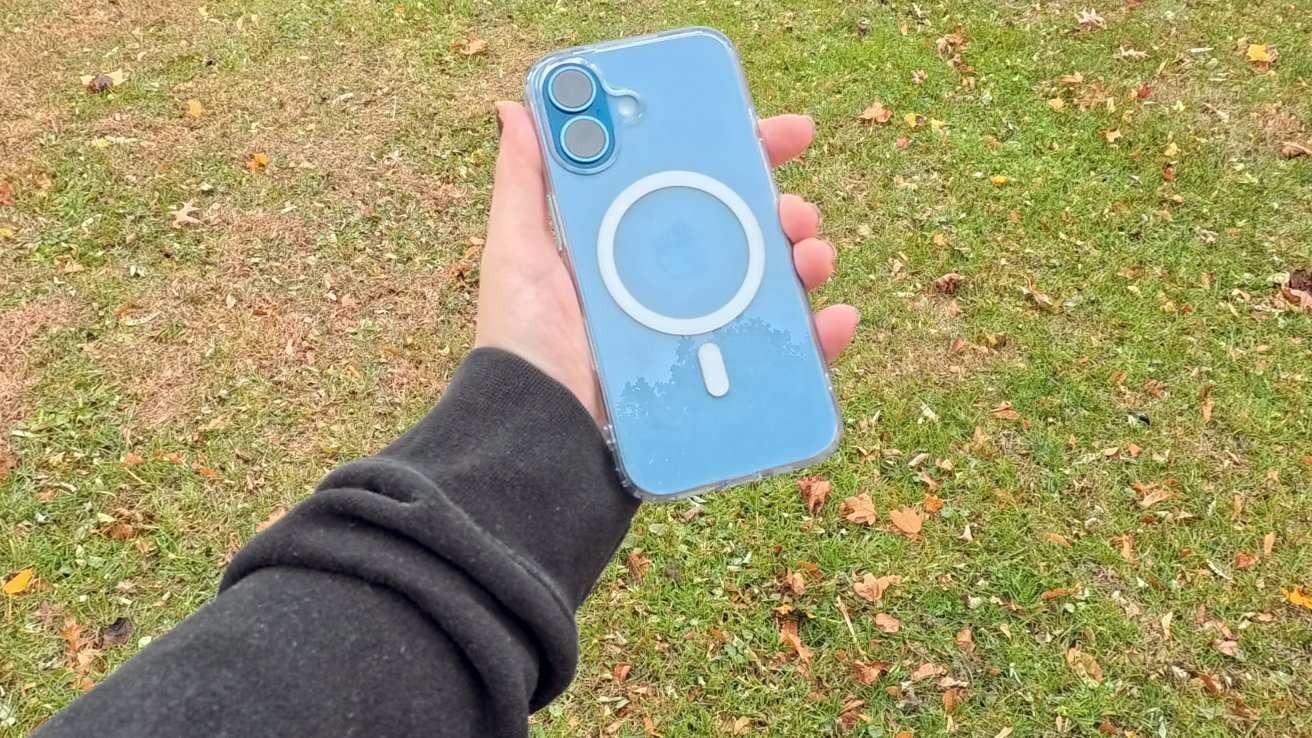 A hand holding a phone with a blue case featuring a circular design, against a grassy background with scattered leaves.
