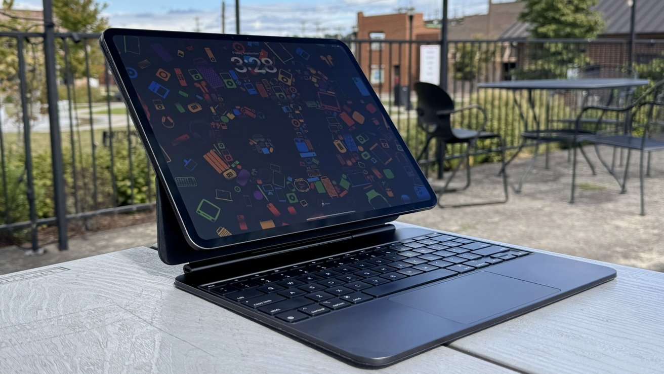 Tablet with colorful screen on keyboard stand outdoors, on a light wood table, with blurred chairs and greenery in the background.