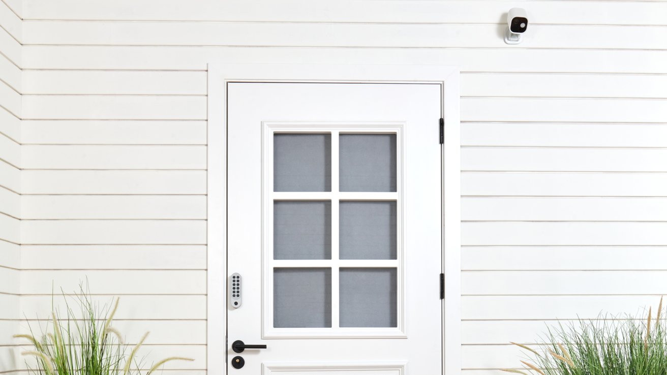White door with six-pane windows set in a horizontal plank wall, electronic keypad lock, and security camera above, flanked by tall grasses.