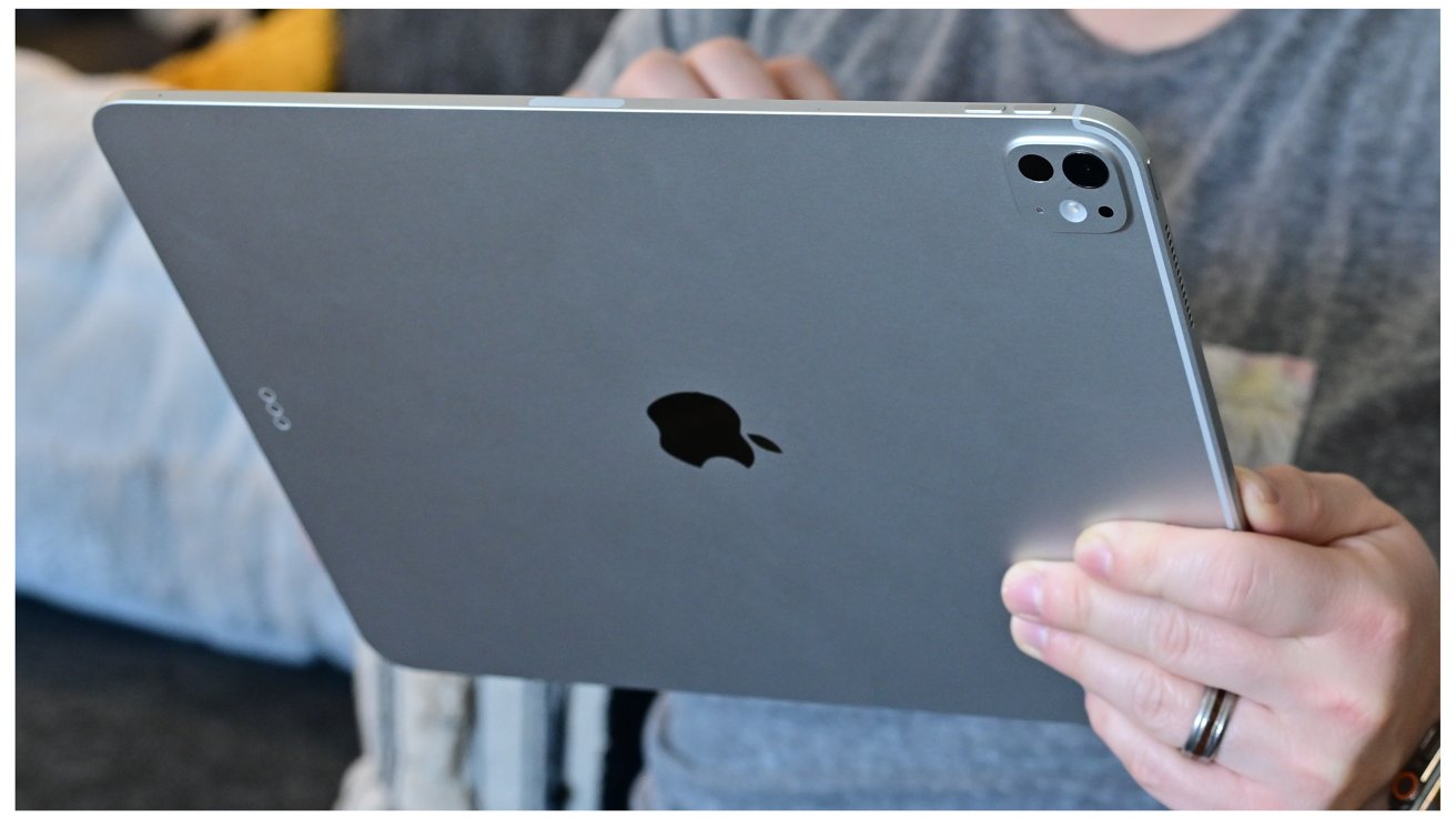 Person holding a large tablet with an Apple logo, featuring dual rear cameras, dressed casually in gray.
