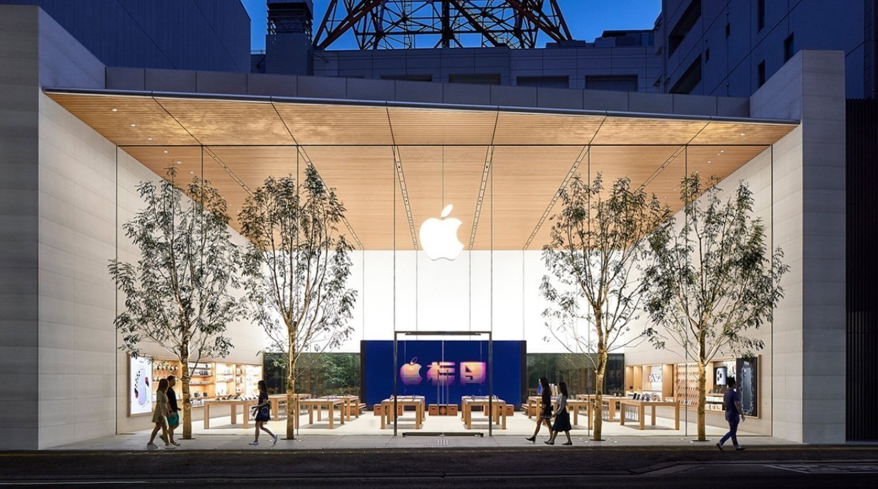 Modern glass-fronted store at dusk, featuring a large illuminated logo, wooden ceiling, interior wooden tables, and trees outside. Several people walk past.