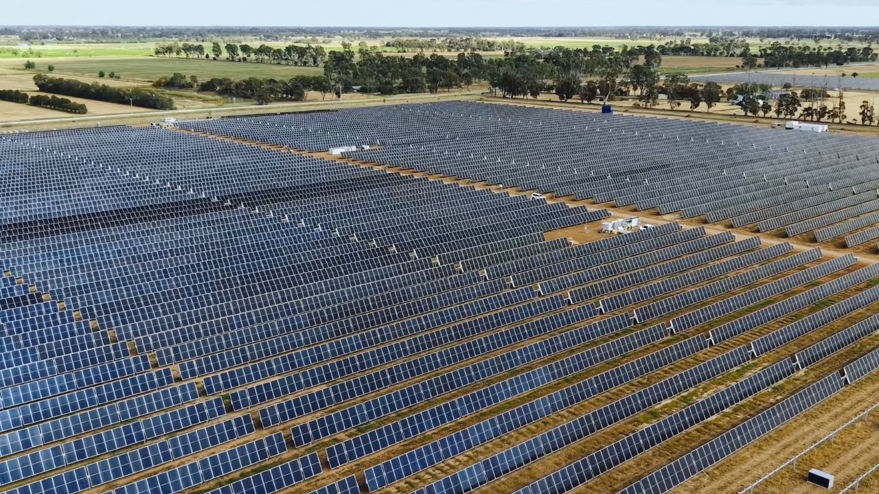Vast solar panel field with rows of dark panels stretching into the distance, surrounded by a grassy landscape and trees under a clear blue sky.