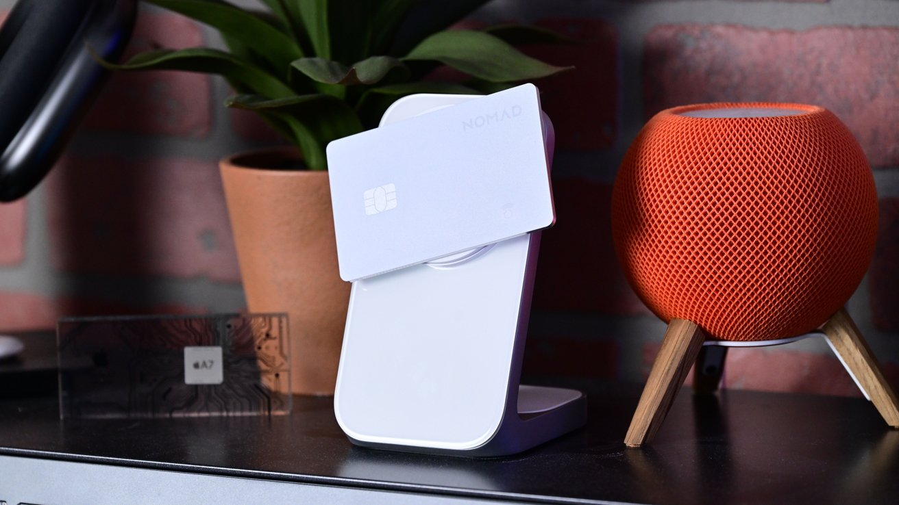 A white card on a wireless charger, next to an orange network speaker, a potted plant and a circuit board display on a black table with a brick wall background.