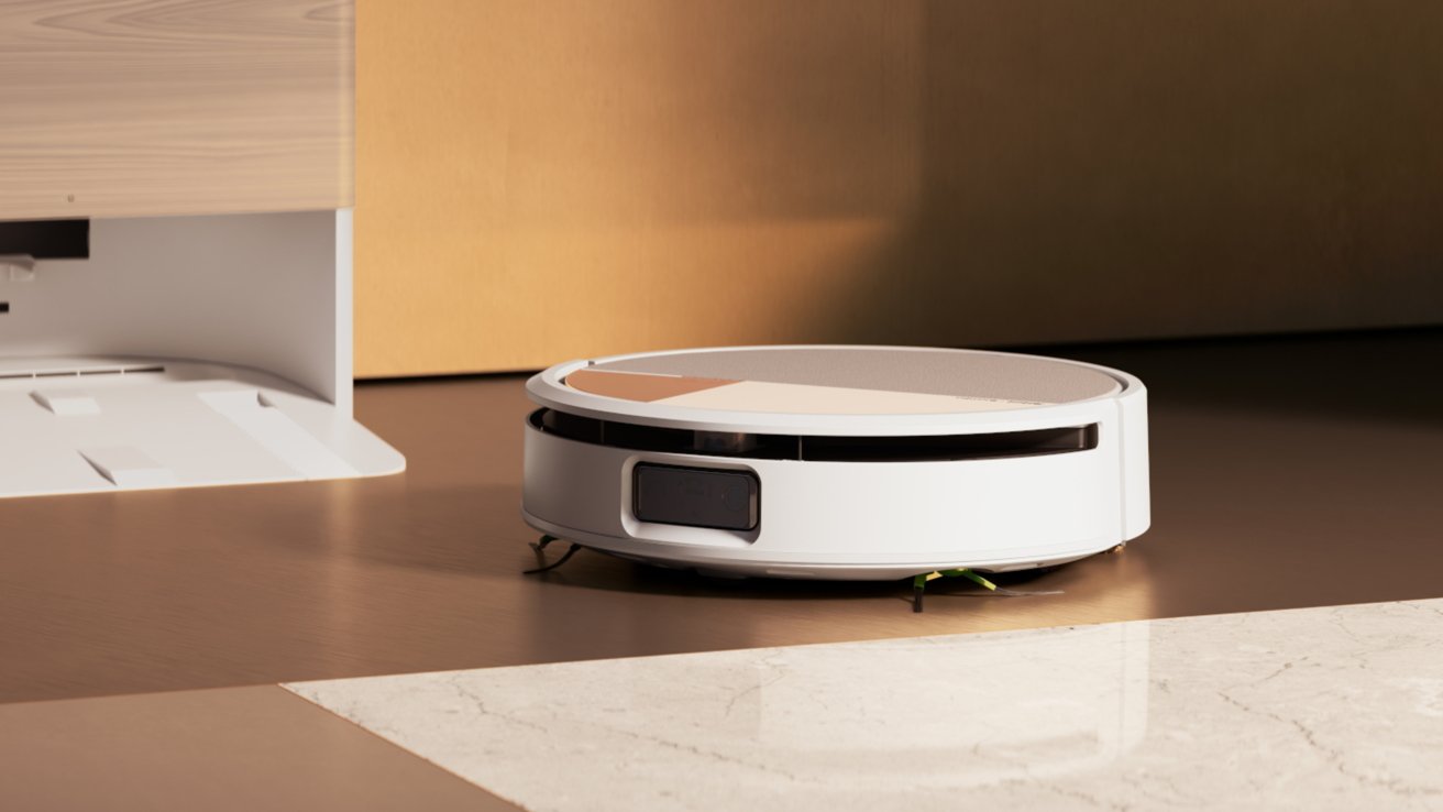 A white robotic vacuum cleaner on a wooden floor near a light-colored wall and charging station, partially on a tiled surface.