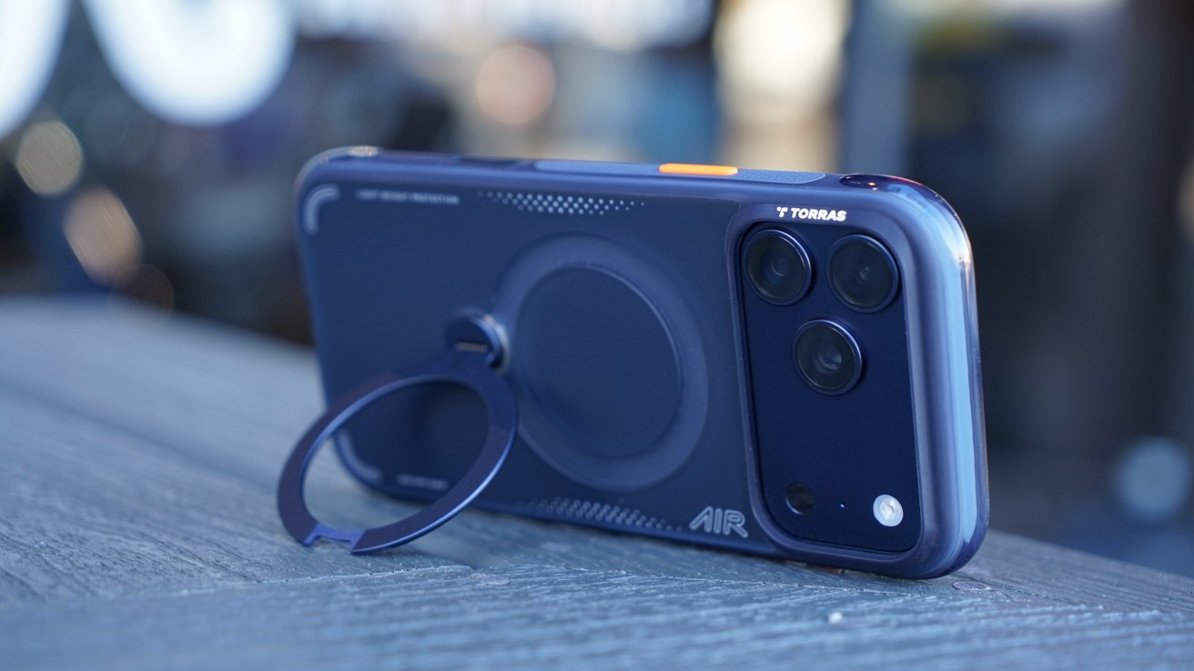 Smartphone with a dark protective case featuring a built-in ring holder, resting on a textured surface. Triple camera setup visible on the back. Blurred background.