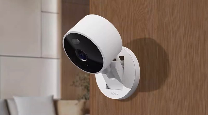A white security camera is mounted on a wooden wall, with a black front facing forward. A blurred living room is in the background.