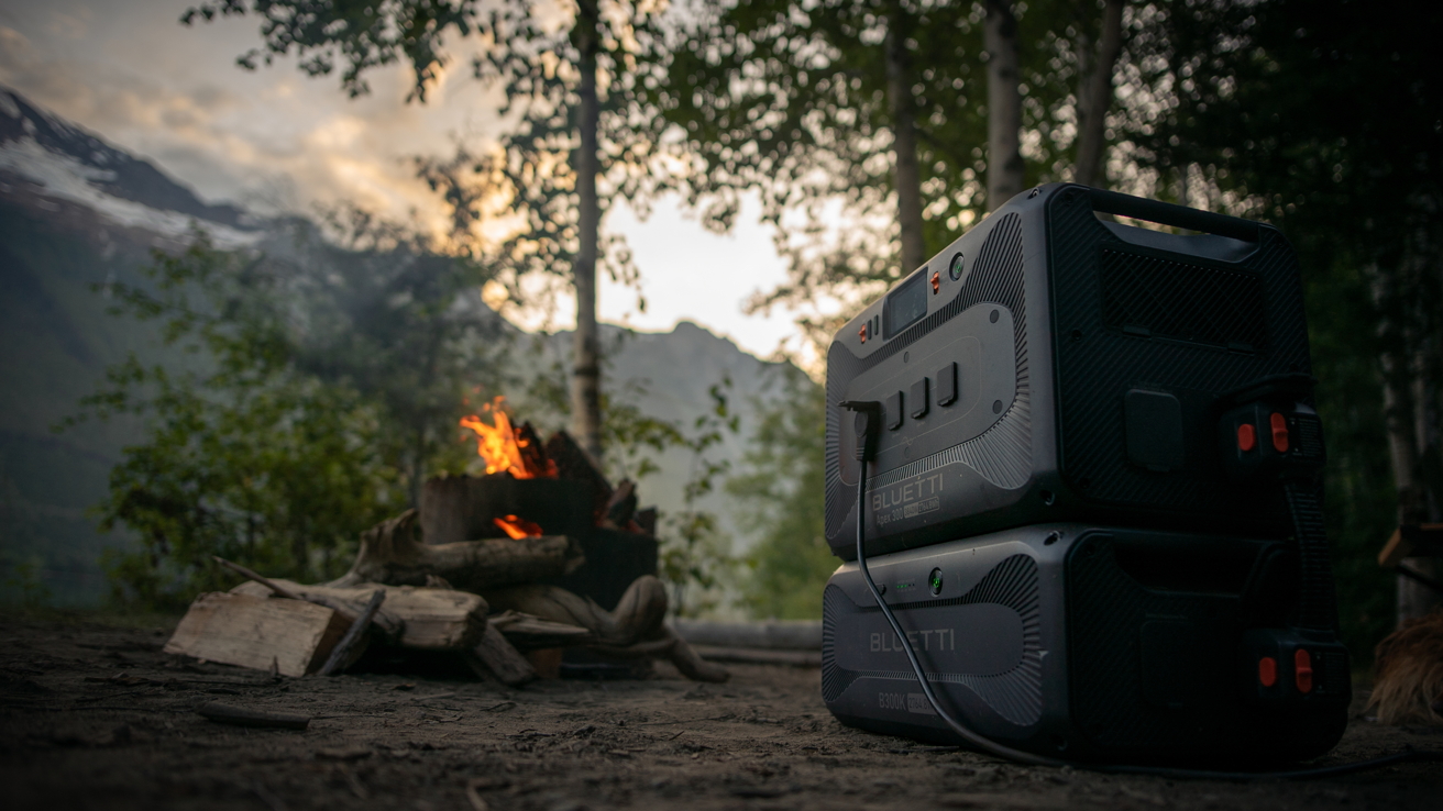 Portable power station stacked near a small campfire in a forest setting with mountainous background and soft evening light.