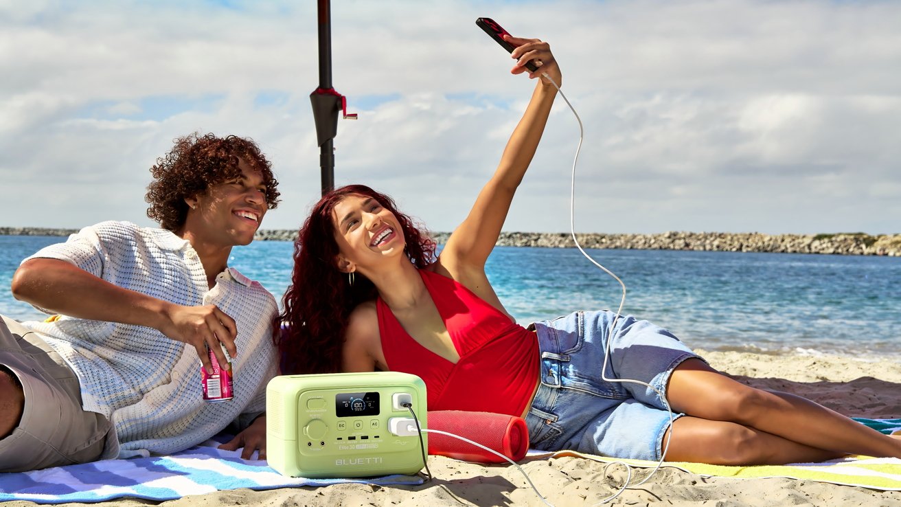 Two people relaxing on a sandy beach, one holding a drink and the other taking a selfie, with a portable power station beside them.