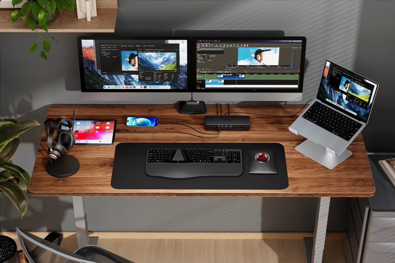 Wooden desk with dual monitors, a laptop on a stand, keyboard, mouse, tablet, smartphone, headphones, plant, and a small tech hub, all in a modern office setup.