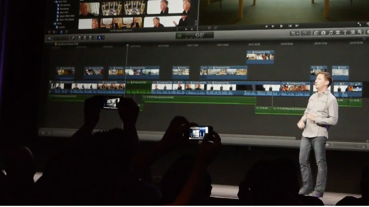 Person presenting a video editing software on a large screen, with audience members holding up phones to record or photograph the presentation.