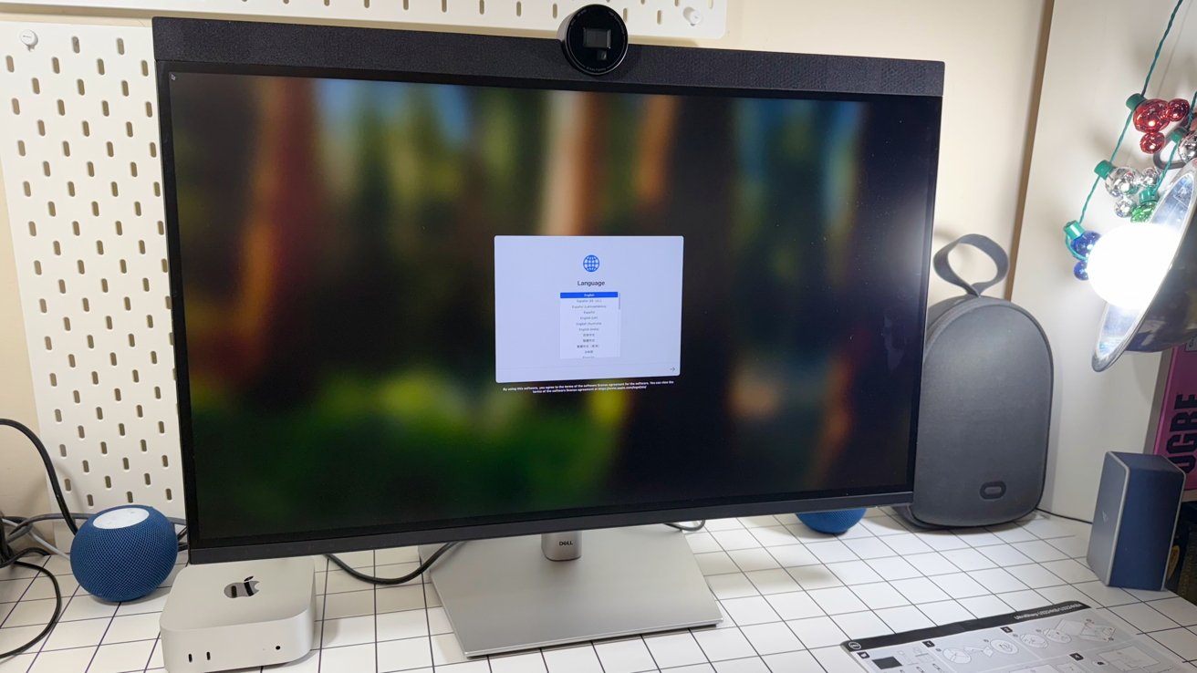 Computer monitor displaying language selection menu, desk cluttered with tech gadgets, peripherals, colorful string lights, and a speaker under bright lighting.
