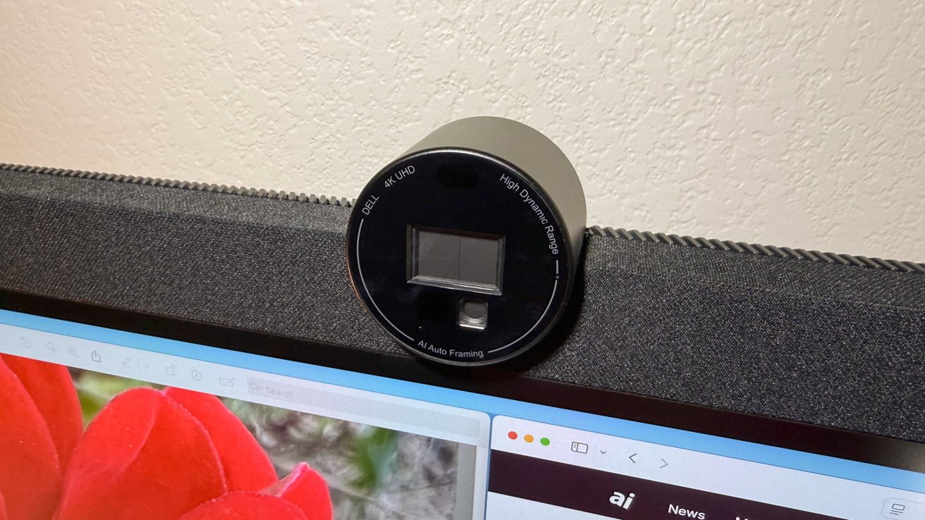 A round camera with automatic AI framing is placed on a fabric surface above a computer screen with a red flower and menu icons.