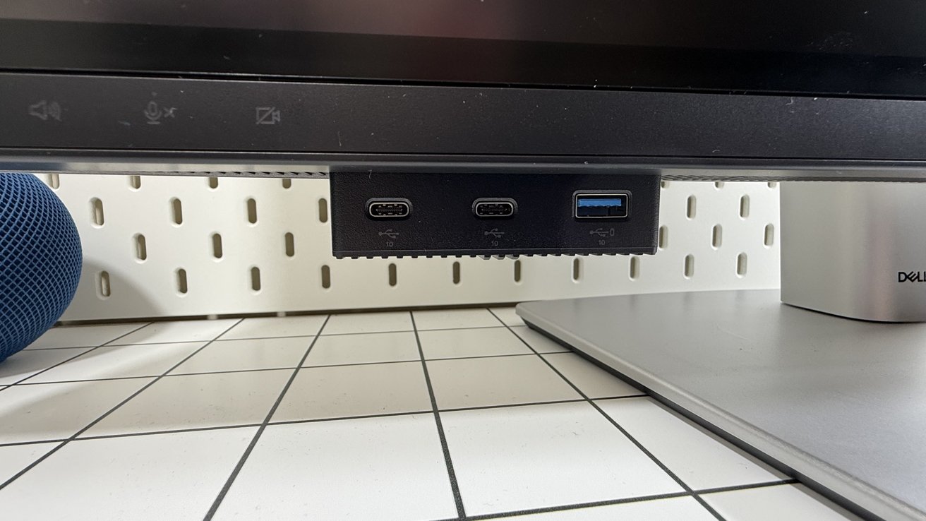 Dell UltraSharp 32 6K monitor with USB-C and USB-A ports on the bottom, a blue speaker and a computer tower on a white table surface with a grid pattern.