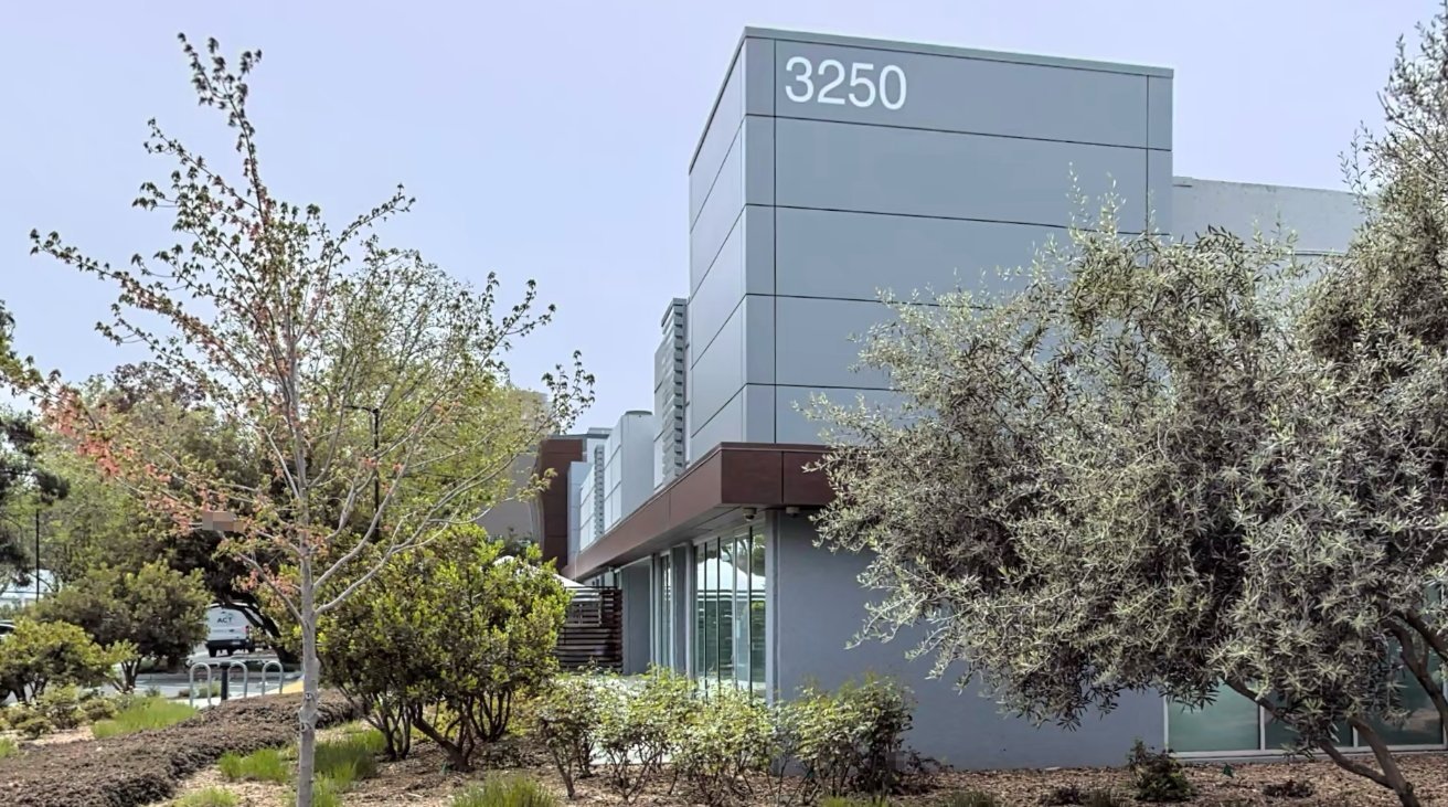 Modern gray building with the number 3250 and large windows, surrounded by trees and plants on a sunny day.