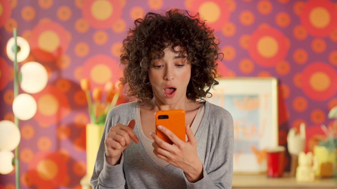 Person with curly hair looks surprised at orange iPhone, bright retro-patterned background with orange and pink circles, desk items, and soft lighting behind.
