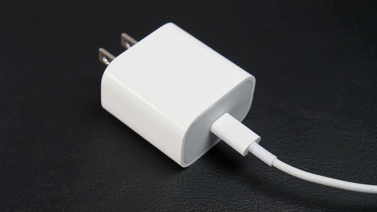 A white rectangular Apple USB-C power adapter with two metal pins and a white cable attached, shown on a black surface.