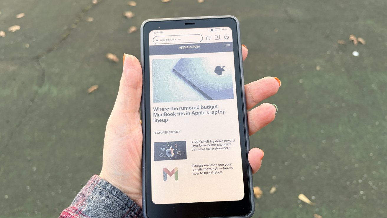 A hand holds a smartphone displaying an AppleInsider webpage about a rumored budget MacBook. The phone is shown against an outdoor pavement background.