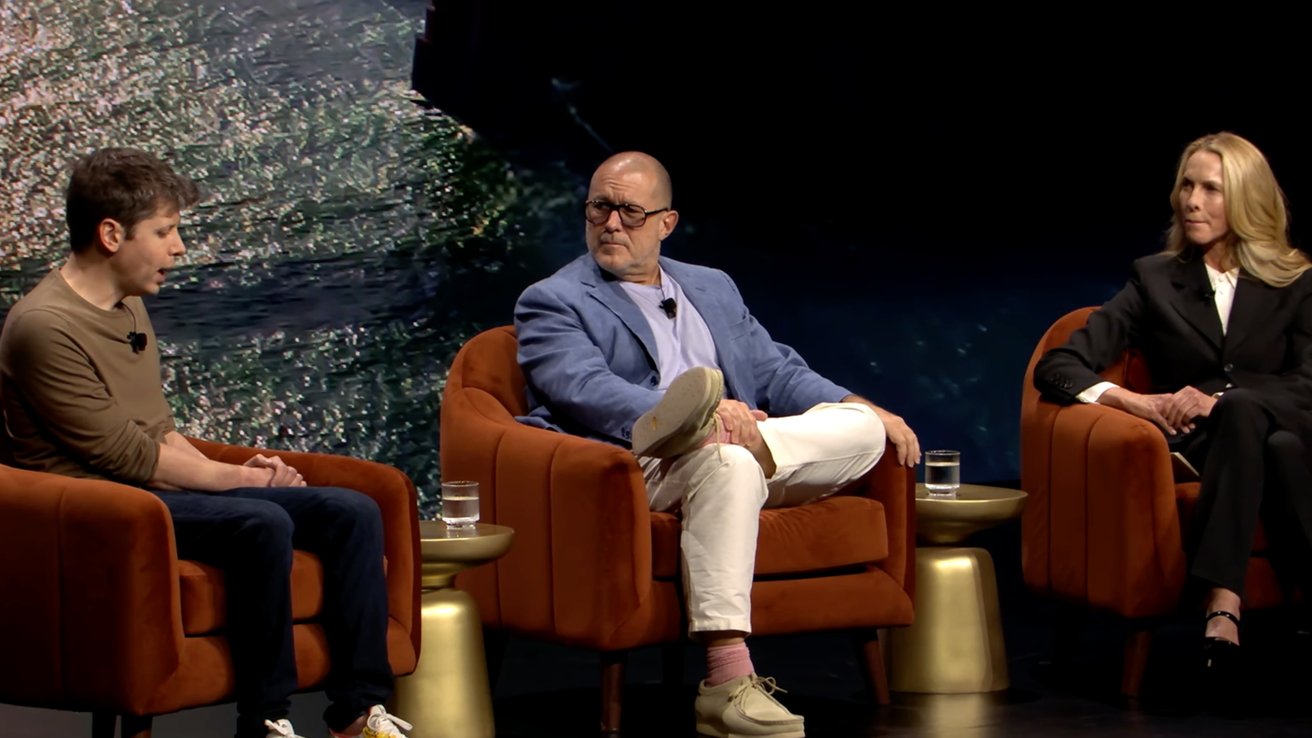 Three people seated on stage in a panel discussion, with brown chairs and small side tables holding glasses of water, against a background of shimmering water.