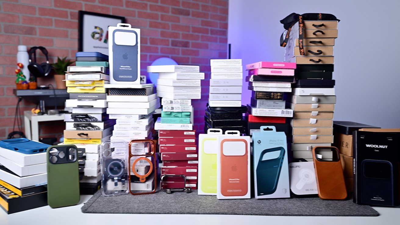 Stacks of smartphone case boxes in various colors and sizes on a table, with a brick wall and shelves in the background.