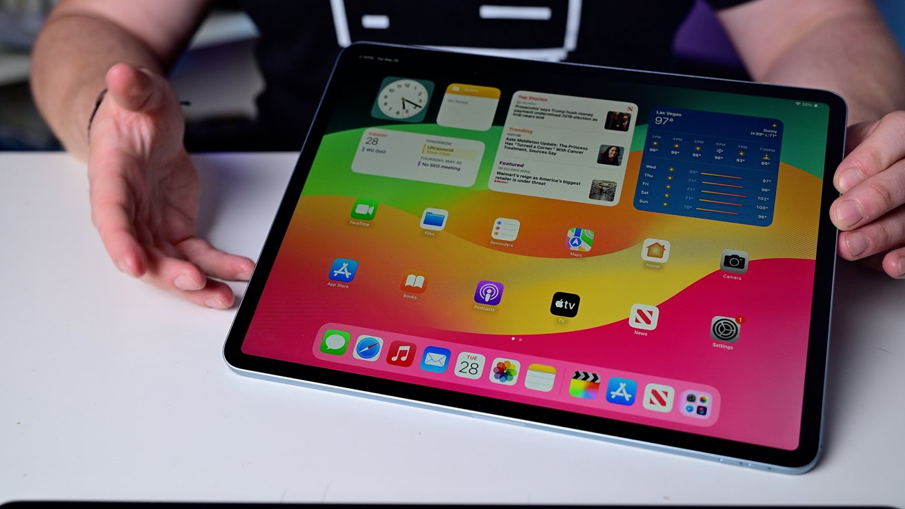 Tablet displaying colorful home screen with various app icons and widgets. Two hands hold the device on a white surface, with screens showing news, calendar, and weather.